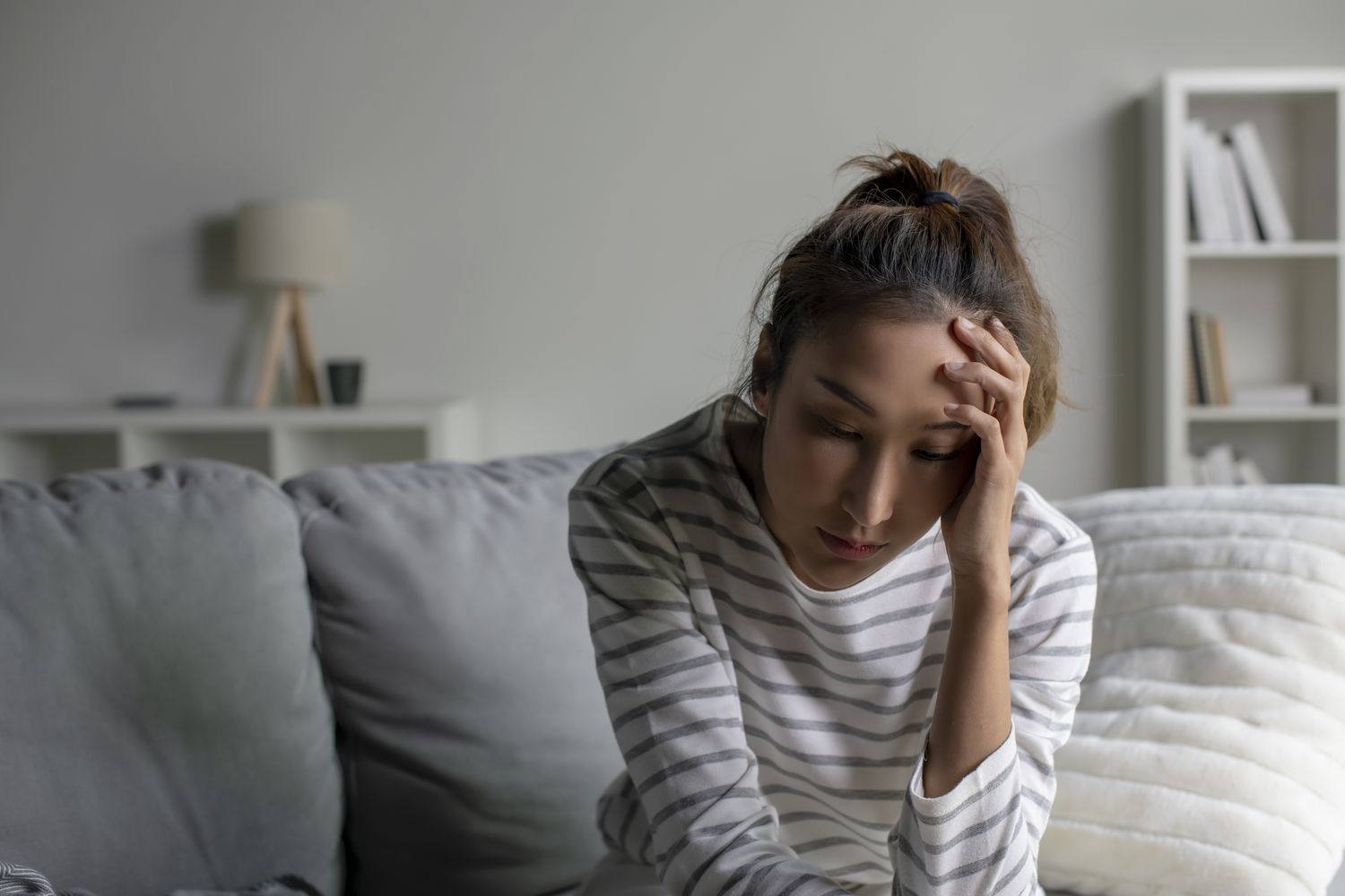 A woman in a striped shirt sitting on a sofa looking tired and stressed with her hand on her forehead.