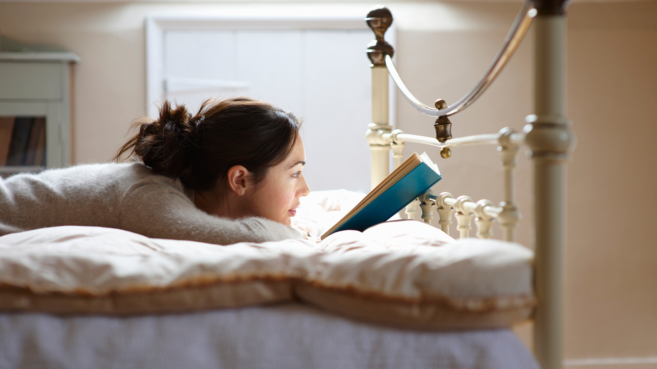 A woman lying in bed reading a book under warm lamp light.