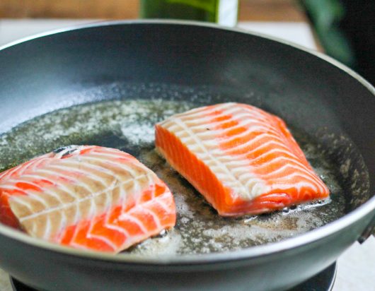 Salmon fillets being seared skin-side down in a hot stainless steel skillet.
