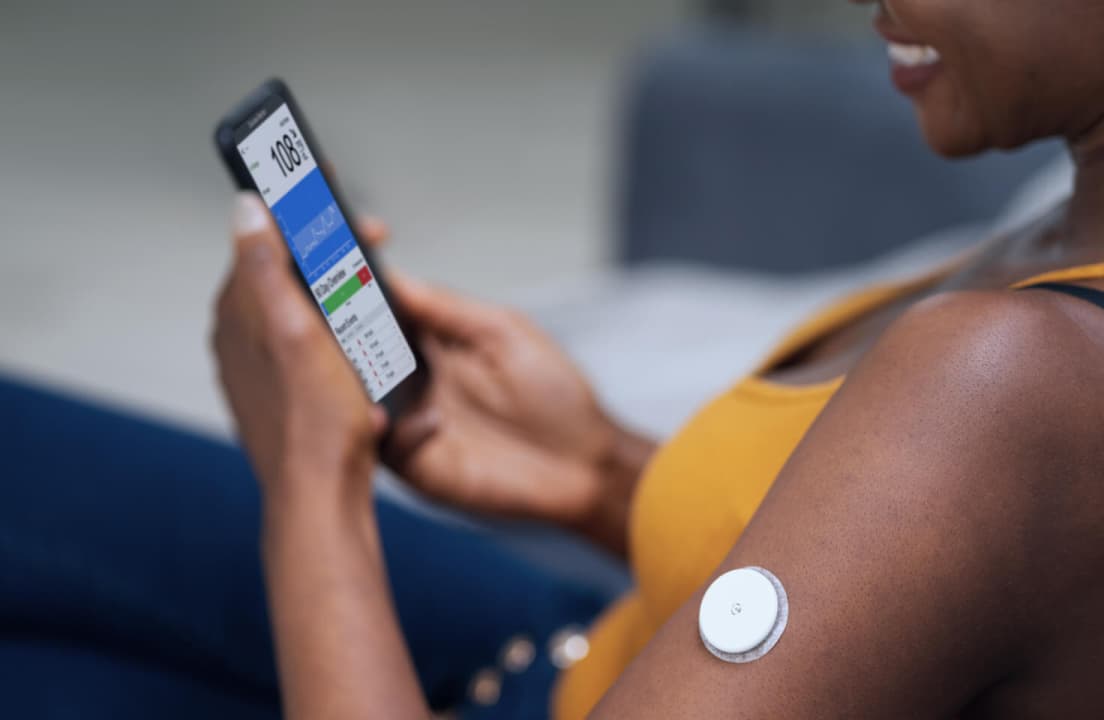 A close-up shot of a small, white circular continuous glucose monitor sensor applied to a person's upper arm.