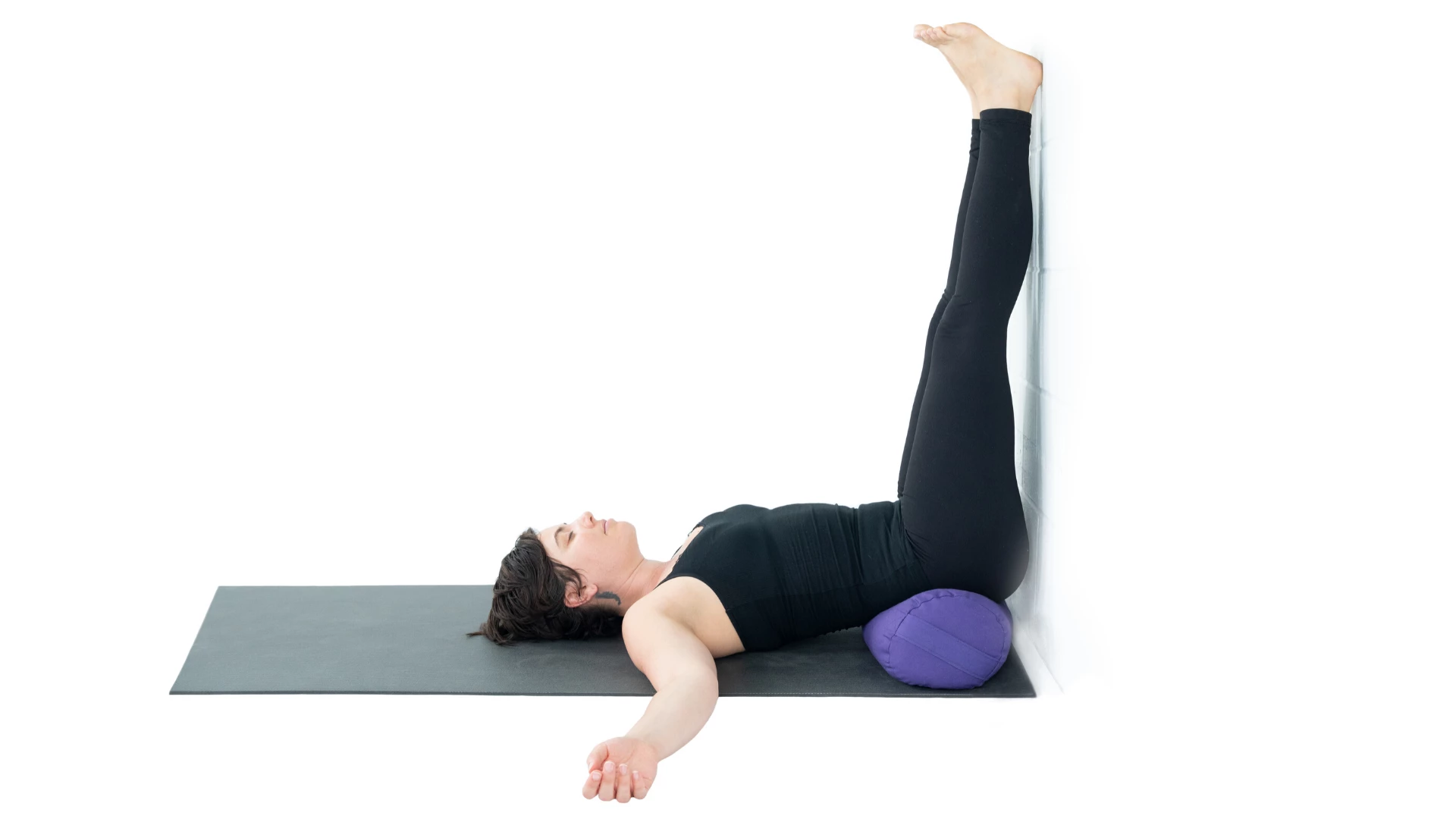 A woman lying on a yoga mat with her legs resting vertically against a white wall.