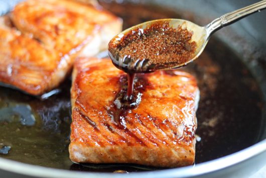 Basting salmon fillets with a thick, glossy glaze using a spoon in a hot pan.