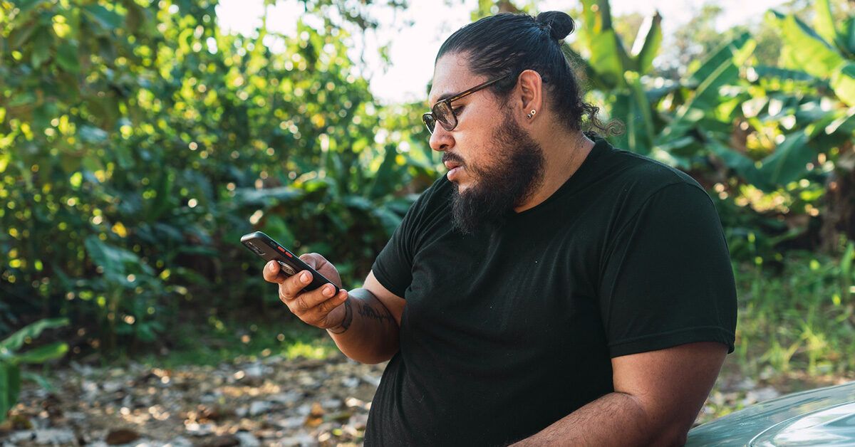 A man in a green outdoor setting looking at his smartphone with a focused expression.