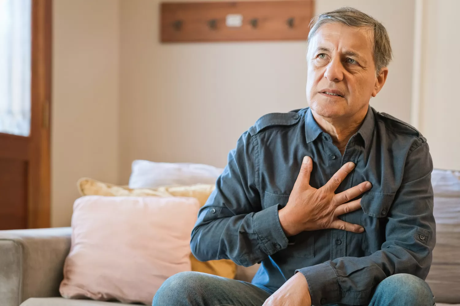 An older man sitting at home holding his hand to his chest in visible discomfort.
