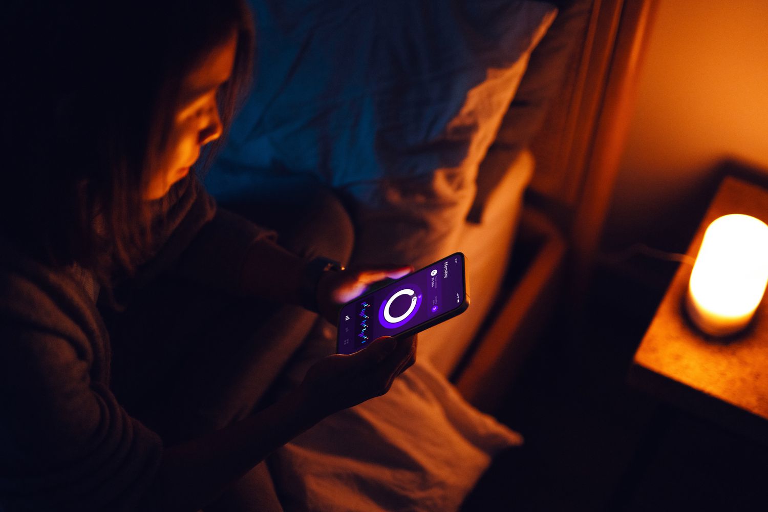 A woman checking a health application on her smartphone while sitting on her bed at night.