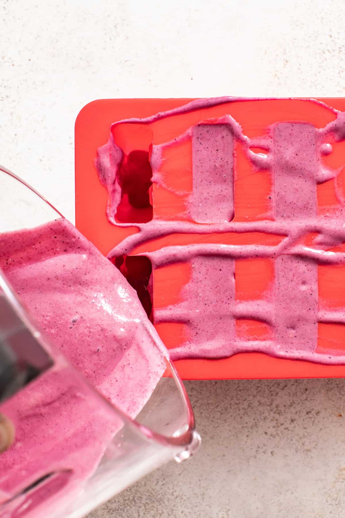 A hand pouring purple berry smoothie mixture into a blue silicone popsicle mold.