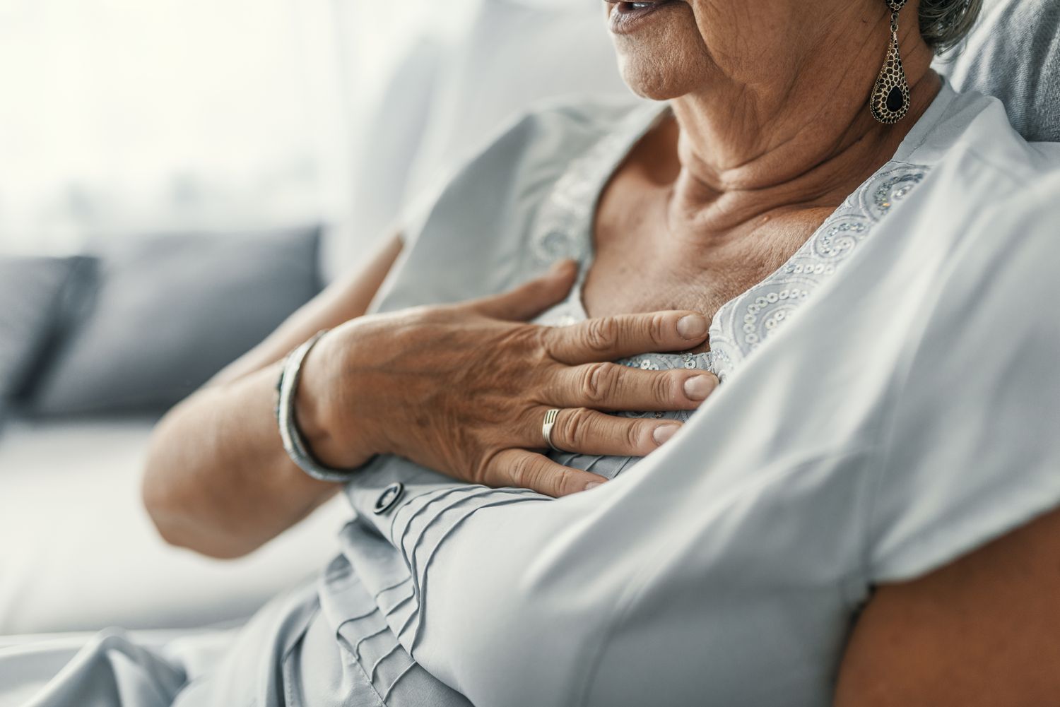 An elderly woman in a light-colored blouse holds her chest, demonstrating signs of physical distress or pain.
