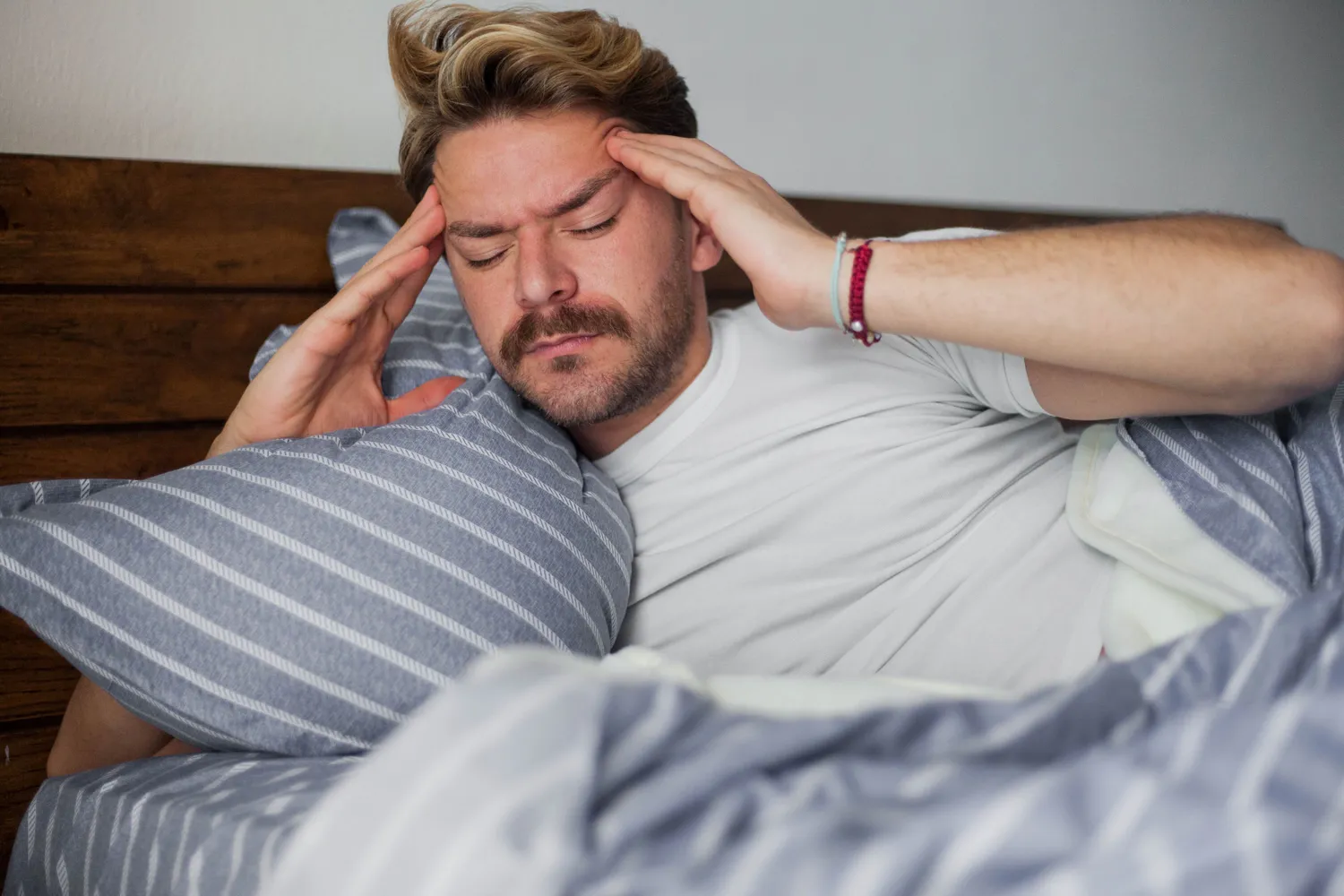 A man lying in bed looking distressed and holding his head in discomfort.