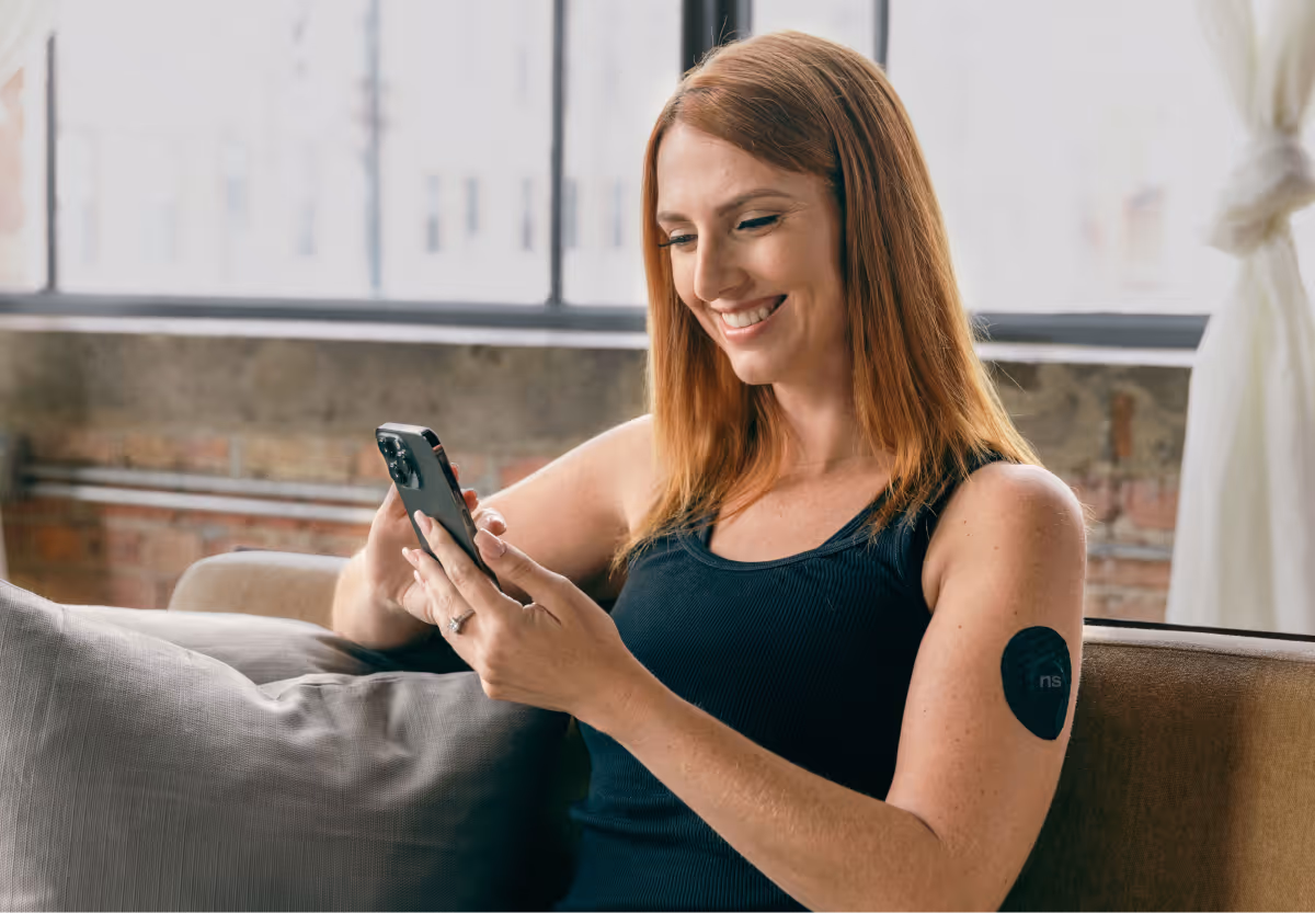 A woman sitting on a couch checking her real-time glucose trends on a smartphone app.