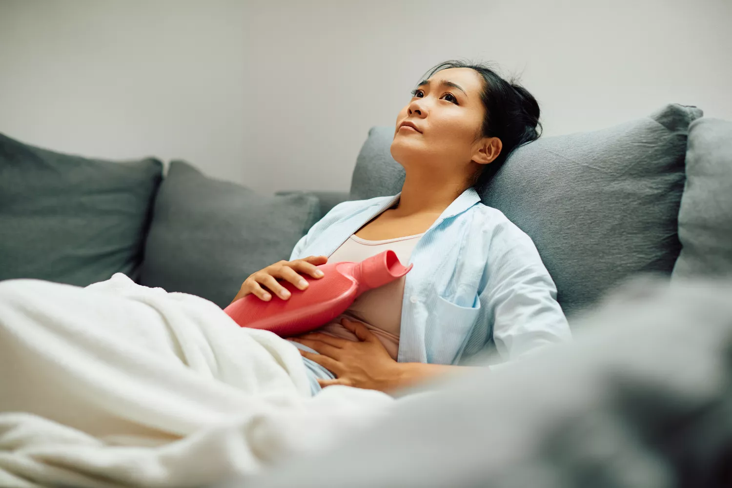 A young woman sitting on a couch expressing discomfort while holding a hot water bottle against her stomach.