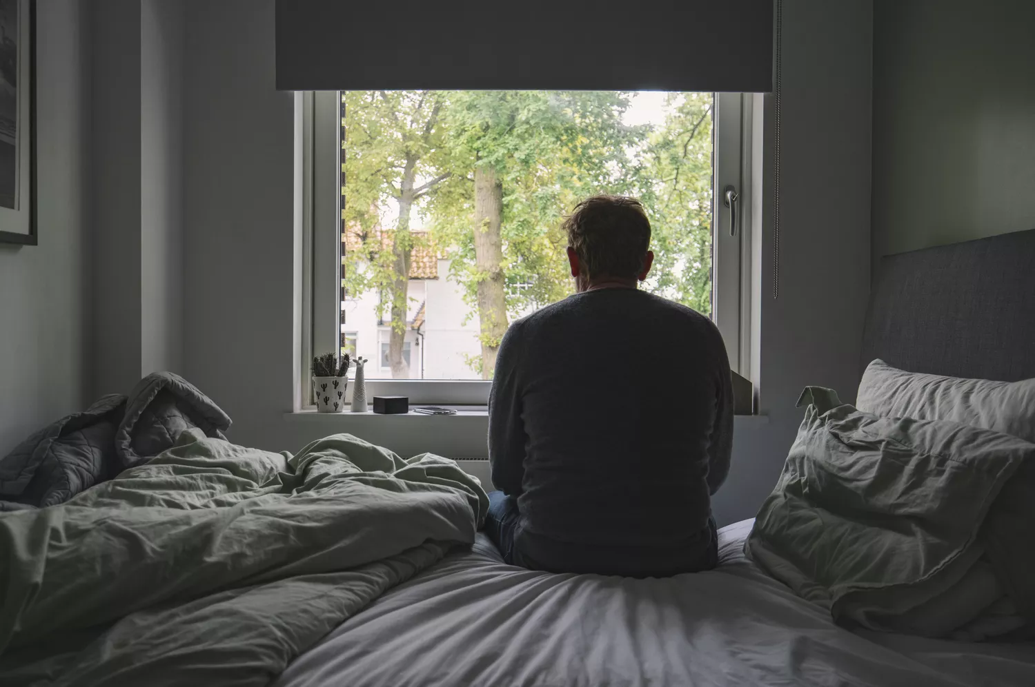 A man sitting on a bed looking out a window, reflecting the emotional aspect of health management.