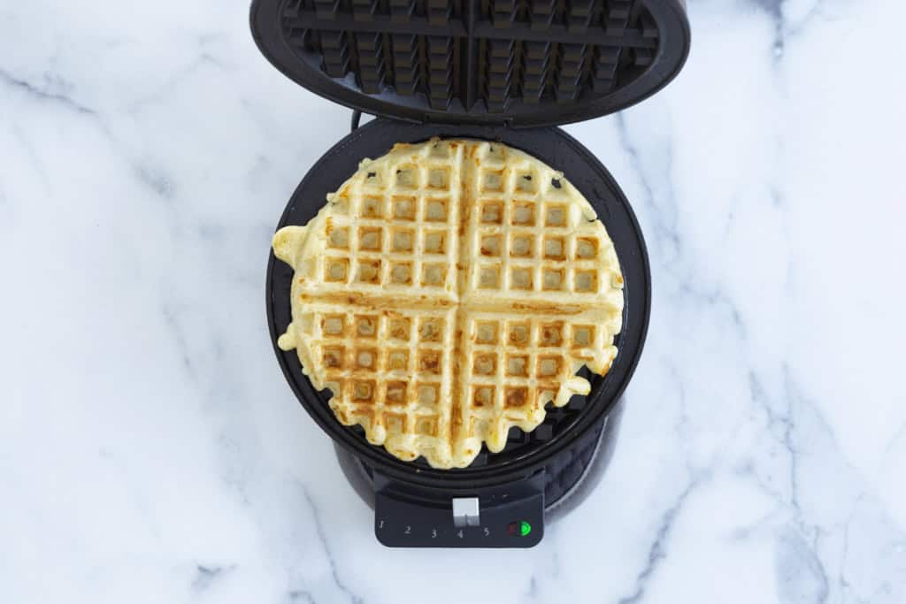 A savory vegetable waffle being cooked in a round waffle iron, showing a crisp golden-brown exterior.