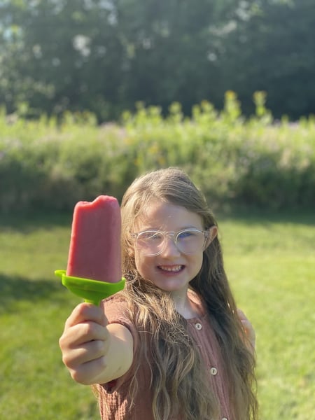 A young child smiling while eating a bright red homemade fruit popsicle.