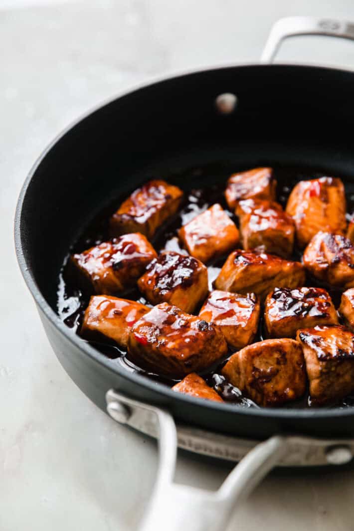 Cubed salmon pieces being glazed and cooked in a dark frying pan.
