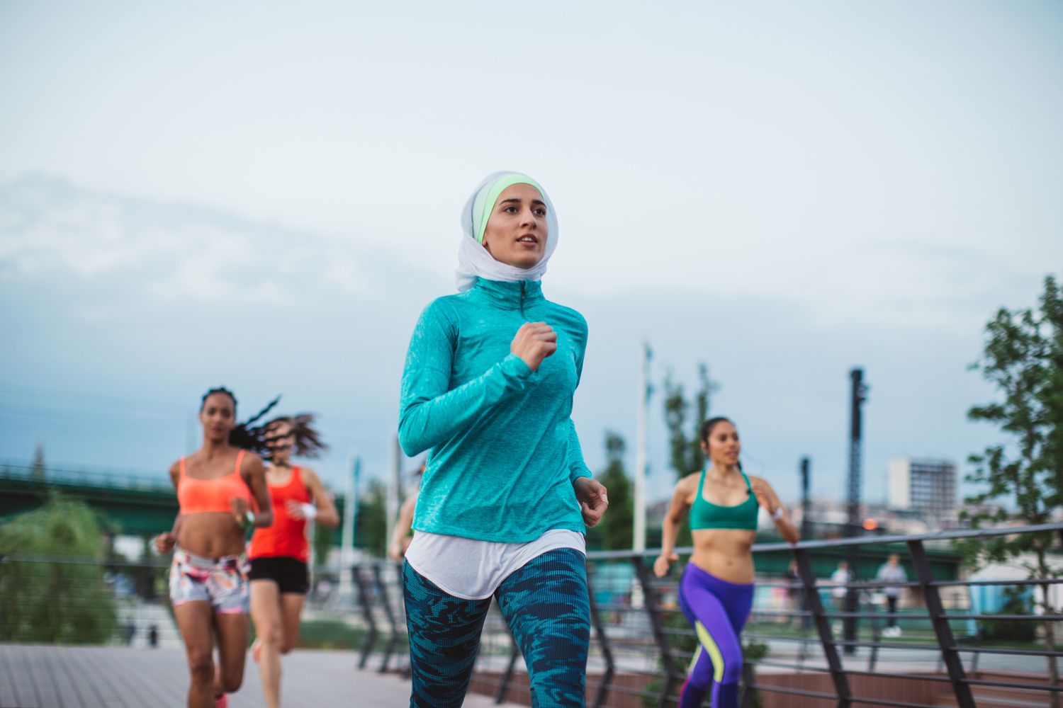 A diverse group of female runners, including a woman in a hijab, running together across an urban bridge.