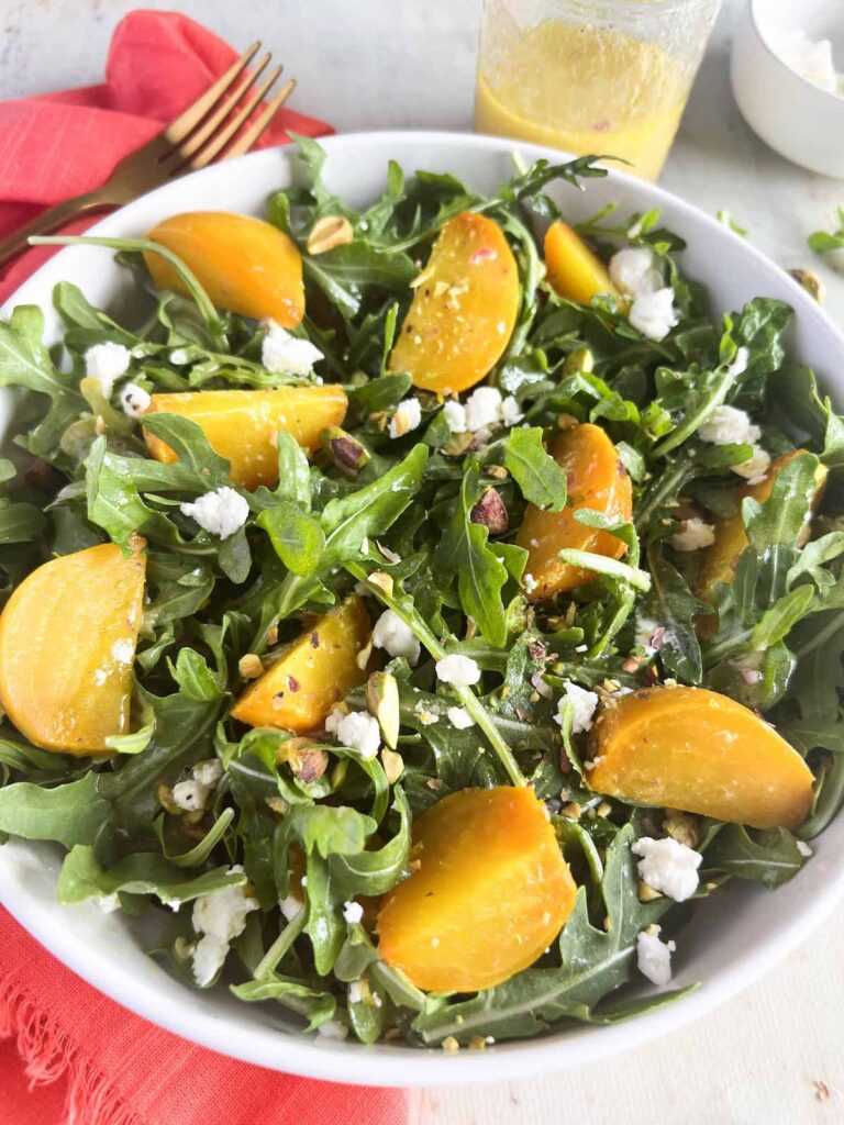 A close-up shot of a golden beet salad with arugula, goat cheese, and crushed pistachios in a white bowl.