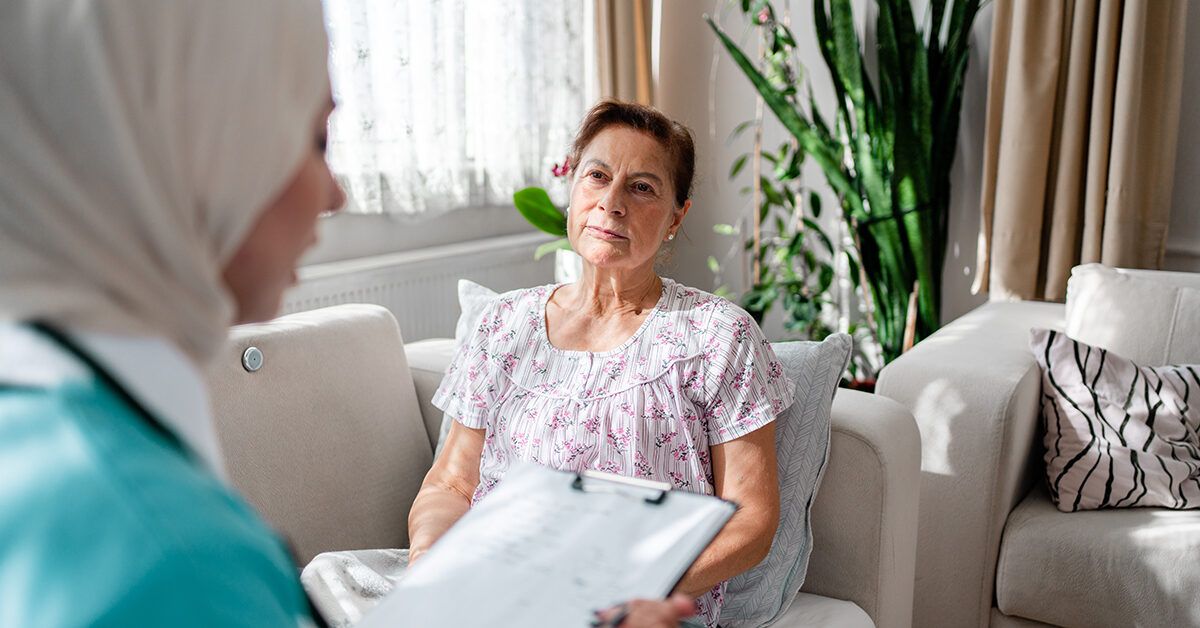 An oncology professional holding a clipboard and talking empathetically to an older patient in a bright room.