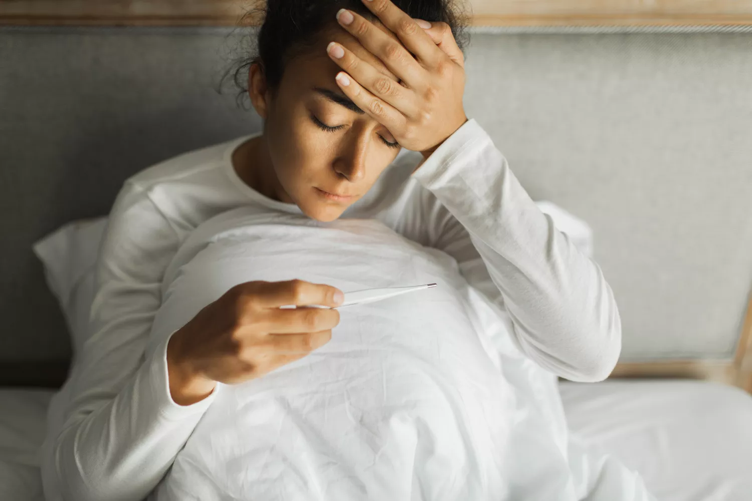 A person resting in bed with a thermometer, appearing unwell.
