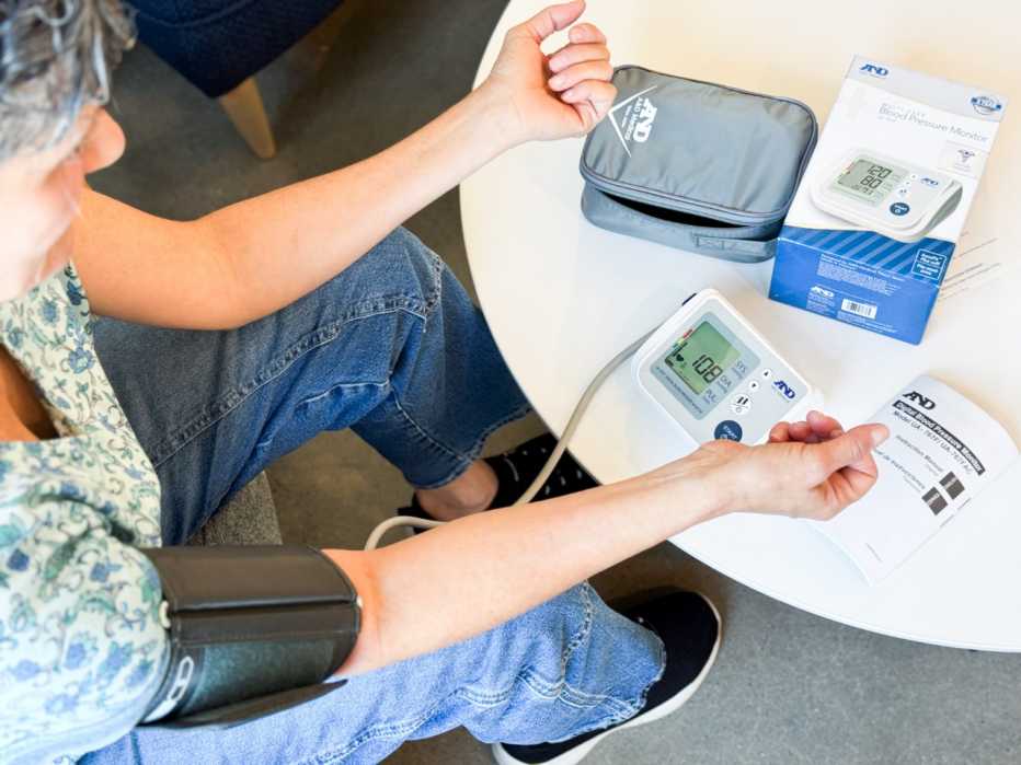 A woman at home using a digital blood pressure monitor with the cuff on her left arm, looking at the device.