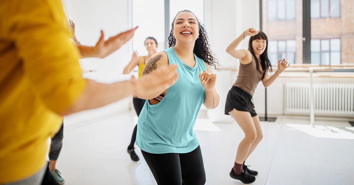A group of women laughing and dancing together in a brightly lit fitness studio.