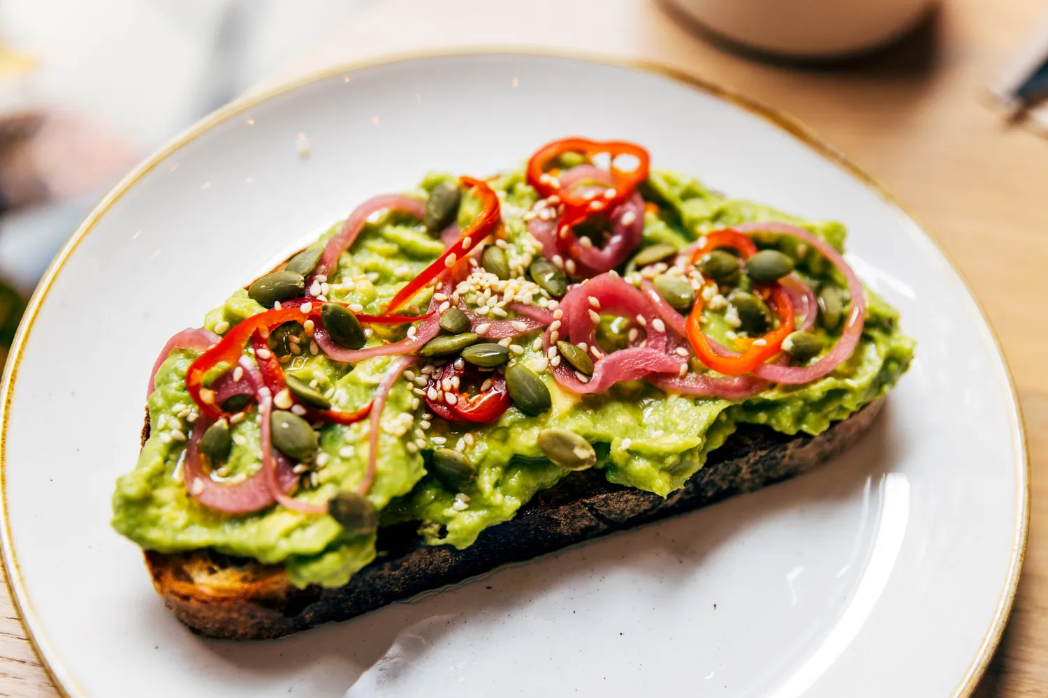 Avocado toast on a white plate topped with peppers, seeds, and pickled onions.