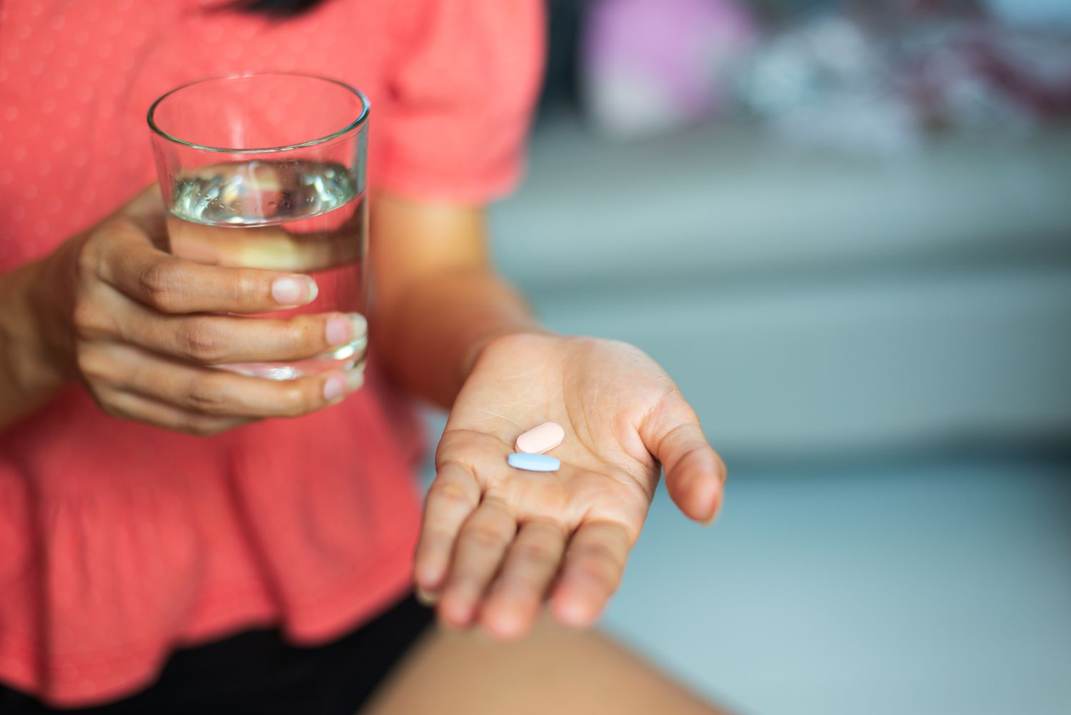 A close-up of a person holding a glass of water and two pills in their palm, representing medication intake.