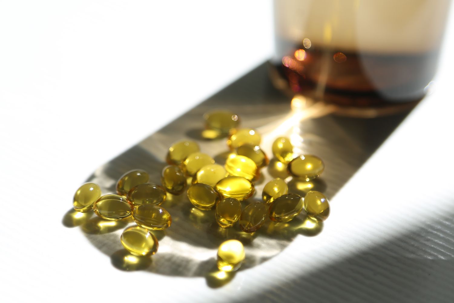 Close-up of golden gel capsules scattered on a surface near a bottle in natural sunlight.