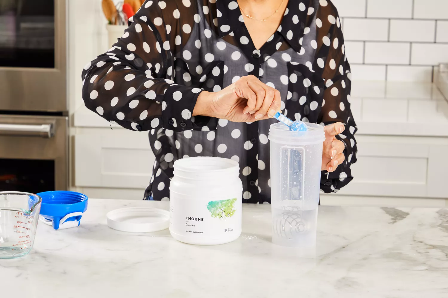 A person adding a scoop of creatine powder into a shaker bottle in a kitchen.