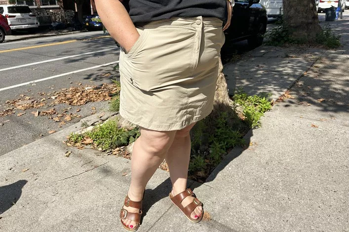 A woman in a beige skirt and black top walking on a sunny city sidewalk.