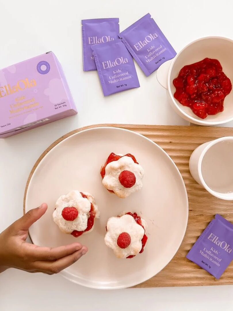 A hand placing a dessert with berries next to EllaOla vitamin sachets.