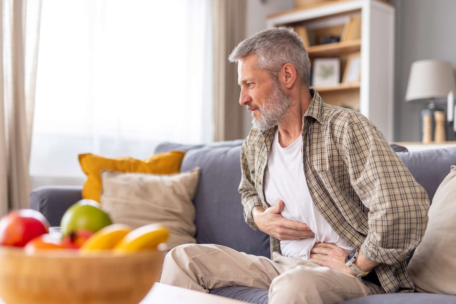 An older man sitting on a couch with a hand on his stomach, looking distressed due to bloating or gas pain.