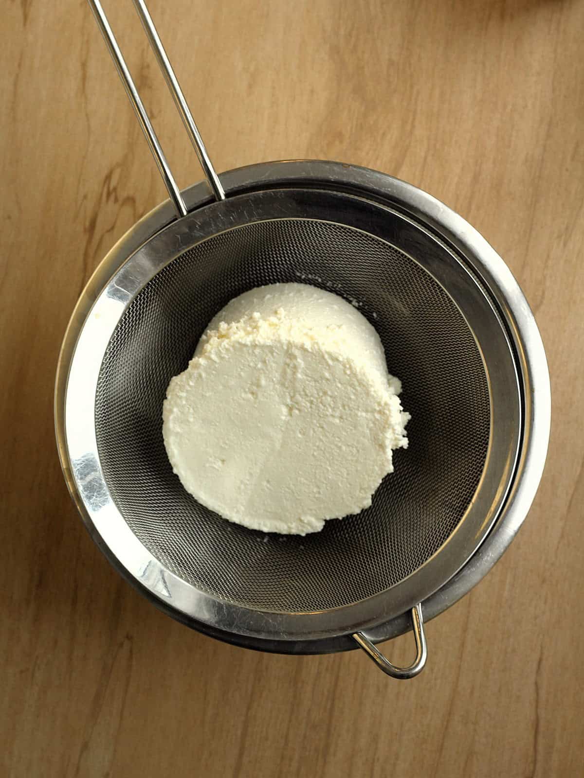 Fresh ricotta cheese draining through a fine mesh sieve over a glass bowl.