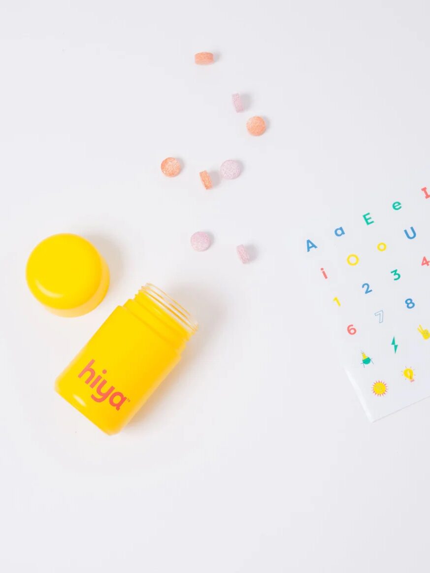 A yellow Hiya multivitamin bottle with several colorful round tablets and a sticker sheet.