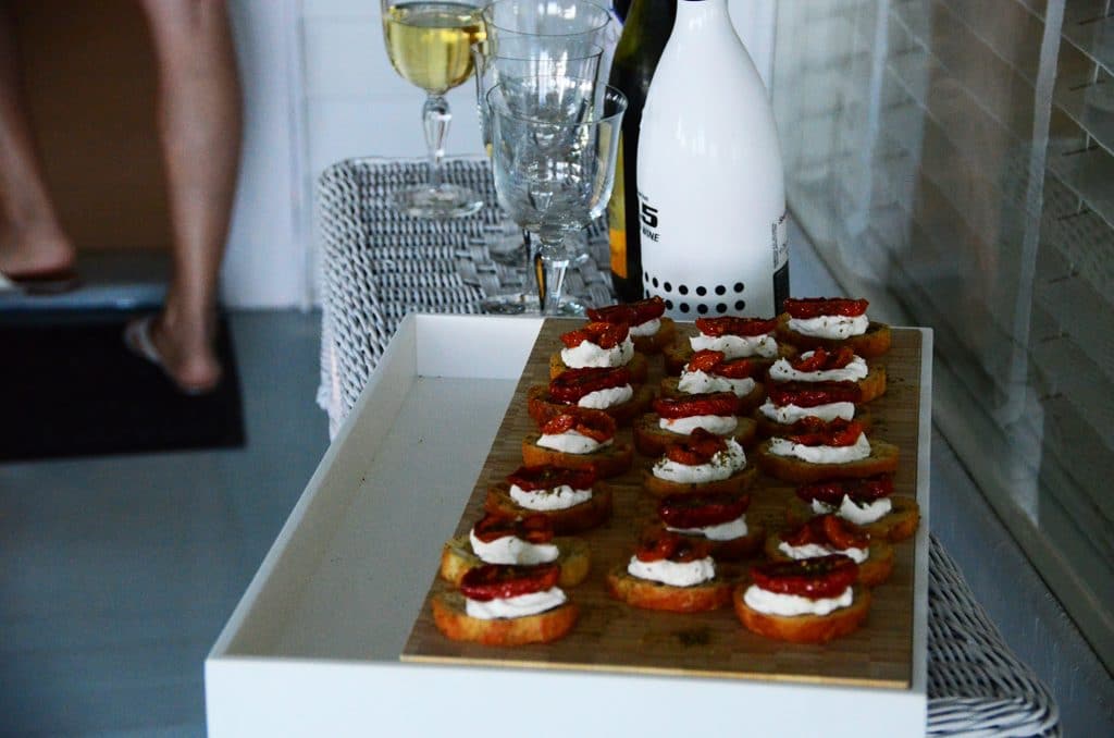 A platter of labneh bruschetta served on a cutting board alongside wine glasses.