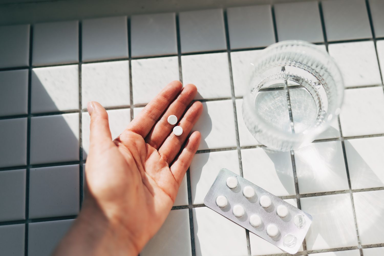 A person holding pharmaceutical pills in their hand next to a glass of water and a blister pack.