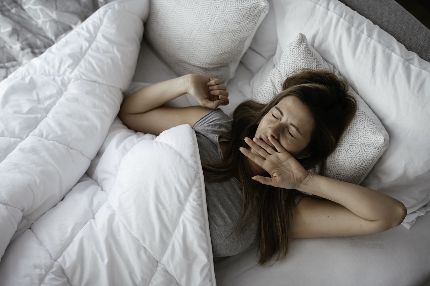 A woman yawning in bed with white linens, looking tired despite the extra hour of sleep.