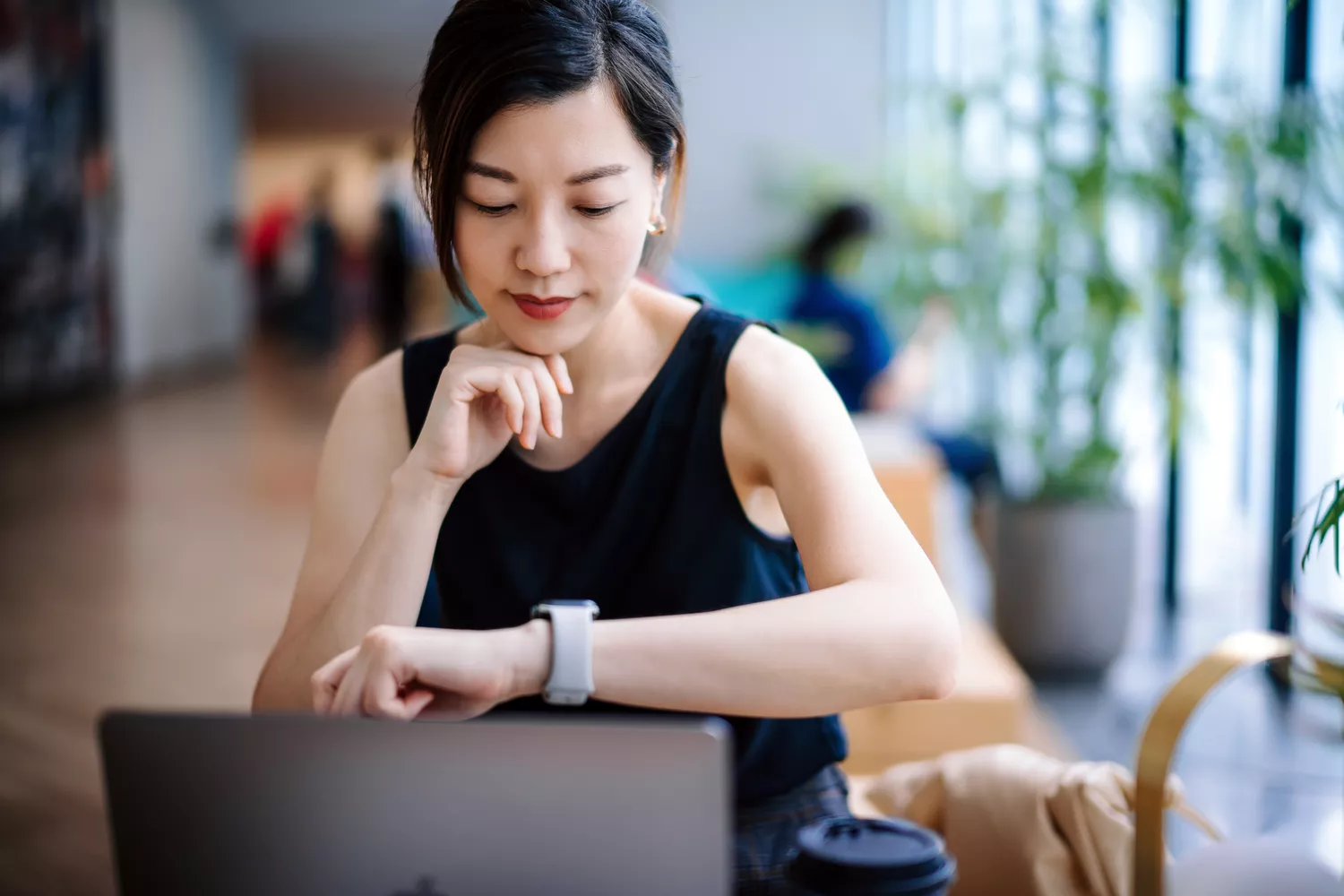 A woman at a desk looking at her watch while working on a laptop, appearing distracted.